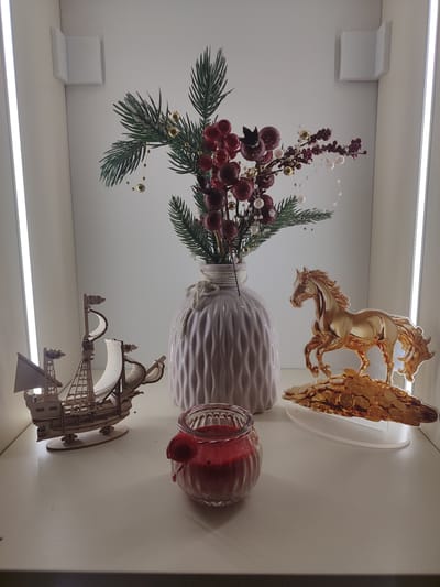 The image displays a still life arrangement on a white shelf, possibly in a display case, with vertical LED lighting on either side. The collection of objects includes a wooden model of a sailing ship on the left, a glass jar containing a red candle in the foreground, and a decorative vase with a festive arrangement of faux pine branches and red berries in the center. To the right of the vase is a gold-colored acrylic figurine of a horse standing on a pile of gold coins. The overall impression is one of decorative, perhaps seasonal, display. The location in the city of Chuy, Kyrgyzstan is not discernible from the image itself.
