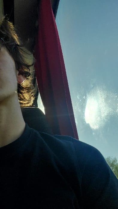 A partial view of an individual is depicted within a vehicle in Coulanges-lès-Nevers, France. The lower left side of the subject's face, neck, and right ear are visible, with dark, wavy hair strongly backlit by natural light. The subject wears a dark, possibly black, crew-neck shirt or sweatshirt. A vertical strip of red fabric, consistent with a curtain or strap, is positioned along the right side of the subject's head. Beyond the subject, a dark vehicle interior component is faintly visible. To the right, a window reveals a clear blue sky. Intense sunlight creates a bright glare and illuminates particles or smudges on the window surface. Faint green foliage is visible through the window in the lower right background.