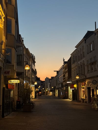 The image depicts a street in Deventer, Netherlands, at dusk. The perspective is from ground level, looking down a cobblestone street lined with buildings. The sky transitions from a clear, pale blue at the top left to a soft orange and yellow gradient near the horizon, indicating the setting sun.
The buildings on either side of the street are multi-story structures with shopfronts at ground level. Some shops have illuminated signs, including "H&M", "TAL4mini" in yellow neon, and another sign with a yellow smiley face. Streetlights are on, casting a warm glow on the street and the lower portions of the buildings.
The overall scene is quiet and peaceful, with no people visible. The presence of bicycles parked near a bin suggests that people use this street for transport. The architecture of the buildings, particularly the facades with their detailed ornamentation, hints at a historic European city. The time of day, twilight, contributes to a serene and slightly moody atmosphere. The weather appears calm and clear, as suggested by the clear sky.
