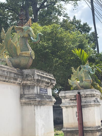 This is a medium shot, vertically oriented photograph taken outdoors in what appears to be Thailand. The scene is dominated by two ornate, green, mythical elephant statues, likely part of a temple or significant structure. These statues are highly detailed, with scales, feathered wings, and elaborate headdresses. The statue in the foreground is larger and more prominently displayed, with its trunk curled upwards. Behind it, another similar statue is visible, positioned higher up and further back.

The statues are mounted on a light-colored, weathered stone structure that forms a wall or building facade. The stone has vertical grooves and appears to be aged, with some discoloration and staining. A red metal beam is affixed horizontally across part of the stone structure in the upper right portion of the image.

In the background, lush green trees with dense foliage fill much of the frame, suggesting a tropical environment. The sky is partly cloudy, with patches of blue visible. The lighting suggests it is daytime, likely late morning or early afternoon, with the sun casting some shadows.

On the stone structure in the mid-ground, a small blue sign with white text is affixed. The visible text, though partially obscured and angled, appears to be in Thai script. The OCR identifies the text as: "เจ้าภาพ สร้างกำแพงถวา 05.สุทธิพร คุณเทียบจิต จิราธิวัดม". This likely indicates a donation or inscription related to the construction or upkeep of the structure. The overall impression is one of cultural and religious significance, with the detailed artistry of the statues contrasting with the weathered, ancient appearance of the stonework.