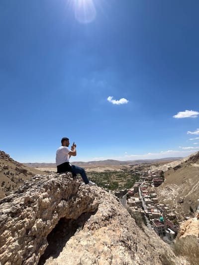 A man is perched on a rocky outcrop overlooking the Syrian city of Ma'loula. He is wearing a white t-shirt, jeans, and white sneakers, and he appears to be taking a picture with his phone. The city of Ma'loula, known for its Aramaic-speaking community, is visible in the background, nestled in the valley below. The sky is blue with a few scattered clouds, and the sun is shining brightly. The landscape is rugged and arid, typical of the mountainous region where Ma'loula is located.