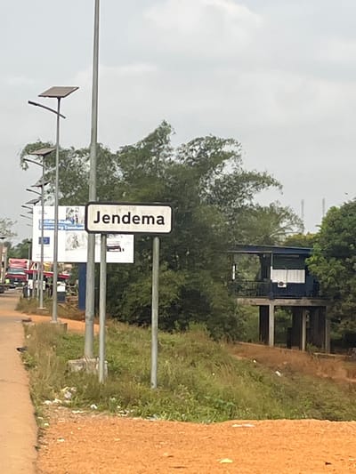 A road sign with "Jendema" in black lettering stands vertically on the side of a road lined with utility poles. In the background, a lush green canopy of trees partially obscures a small, open-air structure with a blue roof and white walls. Other signs and distant vehicles can be seen along the road. The sky is overcast and the roadside vegetation is dense. A dirt embankment appears on the right.