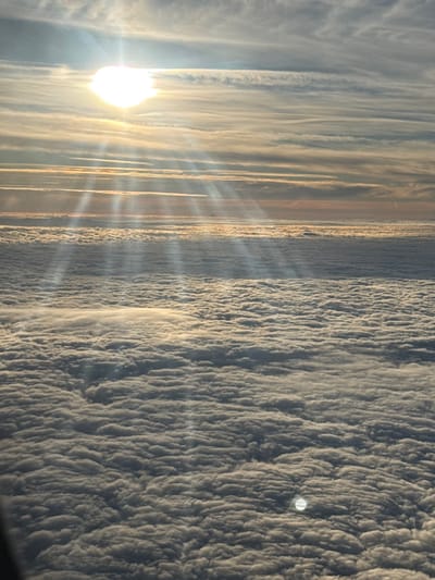 The image displays an aerial perspective of an extensive cloud layer from above. The sun, a bright white orb, is positioned in the upper-left quadrant, emitting distinct crepuscular rays that extend downwards through the atmosphere and illuminate the cloud deck. The sky above the primary cloud formation contains scattered, elongated clouds with hues of pale blue and soft orange near the distant horizon. The predominant feature is a vast, dense, and uniformly textured blanket of low-lying clouds that covers the lower two-thirds of the frame, showing variations in light and shadow due to the direct sunlight. A faint, circular bright spot, possibly a reflection or lens flare, is visible near the bottom-center of the cloud layer. No people, man-made objects, actions, or interactions are discernible. The scene is purely atmospheric. The specific location within the city of Hochheim, Germany, cannot be determined from the visual content provided.