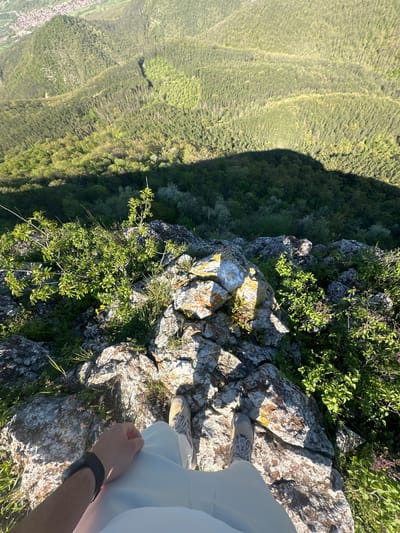 The image shows a person standing on a rocky cliff edge in Bilka, Bulgaria, with a view of a forested valley below. The person is wearing light-colored pants, shoes, and a black watch on their left wrist. The foreground consists of rocks covered in vegetation, while the background reveals a dense forest stretching across the valley. The image is taken from a high vantage point.