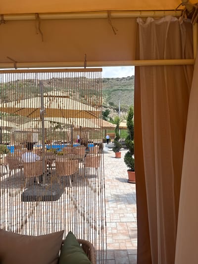 The image depicts an outdoor seating area, possibly a patio or terrace, viewed from under a canopy. In the foreground, a tan fabric canopy is visible, with a sheer white curtain to the left. Beyond the canopy, a view opens up to a landscape of rolling, green hills under a bright sky, suggesting it's daytime with good weather.
The middle ground features a paved area with a repeating geometric pattern, likely stone tiles. Scattered on this paved area are several potted plants, including some with neatly trimmed topiaries and larger shrubs in terracotta pots. A small thatched-roof structure and some outdoor furniture can be observed.
Further into the scene, a series of bamboo blinds or screens create a patterned barrier, partially obscuring a group of people seated at tables. These individuals appear to be engaged in conversation or enjoying a meal, their presence suggesting a social gathering or a dining experience. The overall atmosphere is relaxed and leisurely.
To the far right, a portion of a wicker chair with light-colored cushions is visible, adding to the sense of an inviting outdoor space. The scene evokes a feeling of a pleasant day spent outdoors, likely in a restaurant or resort setting, taking advantage of the scenic surroundings. No text is visible in the image.