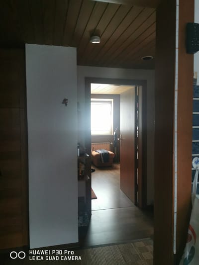 The image shows an interior view of a hallway and an open doorway leading to another room. The hallway features a white textured wall on the left, a wooden ceiling with visible planks, and a wooden door frame on the right. A small decorative item resembling a leaf is affixed to the white wall. The doorway reveals a glimpse into a room with a window, a radiator, and a brown armchair with clothing draped over it. The floor in the hallway appears to be dark wood or laminate, and a woven mat is visible in the foreground. The overall lighting is dim, with a bright light source emanating from the window in the adjacent room. The image includes a watermark at the bottom indicating "HUAWEI P30 Pro LEICA QUAD CAMERA". This image was taken on October 15, 2023, at 14:59. The metadata does not contain specific location information for Bad Oeynhausen, Germany.