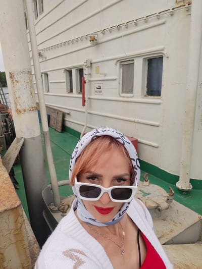 In this medium shot, a woman with reddish-pink hair, wearing a white scarf patterned with a black design and white sunglasses, poses for a selfie on the deck of a white ship. She has on a white cardigan over a red top and a silver necklace. The deck is painted green, and there are various metallic structures and pipes visible. The background shows the white hull of the ship with several rectangular windows, some with their curtains drawn. A red fire extinguisher is mounted on the wall, and a sign with a black pictogram and text in Russian is also visible. The overall lighting suggests it is daytime, and the sky appears overcast, indicating possibly cloudy weather. The scene is set on the water, with a glimpse of green trees on a distant shore, suggesting a location near a river or coastline in Bereg, Russia. The woman's pose and direct gaze into the camera convey a sense of confidence and engagement.
