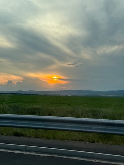 The image captures a scenic view of a rural landscape at sunset, likely in the vicinity of Găgeni, Romania. The foreground is dominated by a dark asphalt road with a concrete curb and a silver metal guardrail running horizontally. Beyond the guardrail, a vast expanse of vibrant green agricultural fields stretches towards a distant line of hazy, undulating hills. The sky above is a dramatic canvas of swirling clouds, tinged with the warm hues of orange and yellow from the setting sun, which is partially obscured by a thick band of clouds. The overall impression is one of serene natural beauty and the tranquil end of a day.