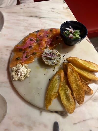 The image displays a close-up, high-angle view of a circular white marble serving platter laden with an assortment of food, resting on a veined light-colored marble tabletop. The specific location within Caminha, Portugal, is not discernible from the visual information provided.
On the platter, the primary subject is a semi-circular arrangement of thin slices of orange-pink smoked salmon, garnished with numerous small green capers and bright pink pickled red onion rings. Adjacent to the salmon, a small, dark blue/black ceramic bowl holds a mix of finely chopped red onion and fresh green herbs, possibly cilantro or parsley, in a liquid. Centrally located is a small mound of finely diced white substance, possibly seafood or fish, topped with thin slices of red onion and scattered black seeds, likely sesame or poppy seeds. In the lower-left quadrant of the platter, a light-colored, piped rosetted dollop of cream or mousse is present. The lower-right portion of the platter features several elongated, golden-brown slices of toasted bread, likely crostini or baguette slices. Scattered black speckles, possibly seasoning, are visible on the white marble platter.
The environment suggests a restaurant setting, indicated by the polished marble table, the formal presentation of food, and the partial view of a red upholstered seat or booth in the background on the right. A portion of a white ceramic bowl is visible on the far left, and a metallic utensil handle is partially visible at the bottom center. No individuals are clearly depicted, though a white fabric, possibly clothing, is partially visible at the top left. The lighting is warm and artificial.
