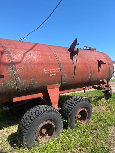This is a medium shot, eye-level, outdoor photograph taken on a sunny day in Laprakasy, Russia. The main subject is a large, weathered, red metal tank trailer positioned on grassy terrain under a clear blue sky. The trailer has two large, treaded tires, and a red, cylindrical tank with visible rust and lichen growth. Cyrillic text is painted on the side of the tank, reading "ВО ВРЕМЯ РАБОТЫ МАШИН БЛИЖЕ 15 М НАХОДИТЬСЯ ОПАСНО," which translates to "During machine operation, stay within 15 meters is dangerous." Below this text, a smaller label indicates "0,37 МП." The trailer also features various metal attachments, including a hinged lid and a pipe. A dark wire is suspended in the sky above the tank, secured to a metal bracket on the tank's surface. The environment consists of green grass, some dry vegetation, and a hint of a white building in the far right background. No people are visible in the image.