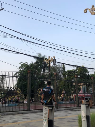 The image shows an outdoor scene, likely a street or public area, with a fence in the foreground and a person walking. The person is wearing a dark t-shirt with blue and yellow accents and text that appears to say "#IMBELIEVEINME" and "@INDONESIAMEDIA". They are holding a phone in their right hand. Behind the fence, there are trees and what looks like a building or structure. Overhead, there are numerous electrical wires against a clear, light blue sky, suggesting daylight. The lighting indicates it is likely daytime, possibly late afternoon given the soft light. The overall scene appears to be a casual, everyday moment. The ground in the foreground is paved with a patterned walkway.