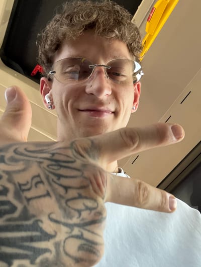 A close-up, selfie-style shot shows a person's face and hand. The person, wearing rimless sunglasses and white earbuds, smiles broadly and holds up a hand with intricate black tattoos. The background features a yellow handle and a dark panel, possibly from a bus or train. The image has a warm, neutral tone. There is no discernible information in the image that can be used to ascertain a location within the city of Navalcarnero, Spain.