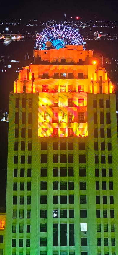 This image is a vertical, high-angle, nighttime shot of a brightly lit tall building. The building is illuminated with warm orange and yellow lights, with some sections also featuring green lighting on the lower floors. The upper portion of the building is topped with a large, illuminated Ferris wheel-like structure, displaying blue and purple lights in a geometric pattern. The windows of the building are visible, with some appearing dark and others lit from within. In the background, a cityscape with numerous distant lights suggests an urban environment at night. The overall scene is vibrant and artificial, dominated by the building's illumination and the colorful structure on its roof.
