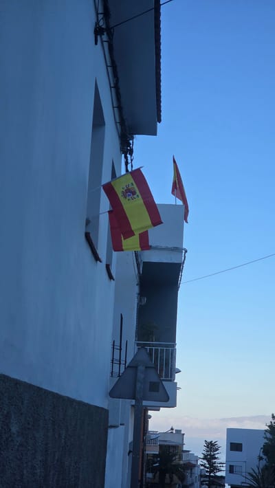 The media file captures a view of buildings against a clear blue sky, suggesting it is daytime. Two Spanish flags are prominently displayed, attached to poles and waving in the breeze. One flag is closer to the foreground, appearing slightly larger and more detailed, while another is further in the background. The flags are the national flag of Spain, featuring red and yellow horizontal bands with a coat of arms on the left side.
The scene shows the exteriors of buildings, with architectural details such as white walls, a tiled roof, and what appears to be an opening or window on the closest building. Electrical wires are visible, strung across the top of the building in the foreground. In the background, other buildings and structures can be seen, some with windows and balconies, and palm trees suggest a warm climate. A peculiar, grey, metallic object with a triangular shape is mounted on a pole, resembling a weather vane or a decorative element. The overall impression is of an outdoor urban or semi-urban environment. The lighting suggests it is either early morning or late afternoon due to the softness of the light and the deep blue of the sky. The location context indicates this is Guía de Isora, Spain. There is no visible text in the media file.