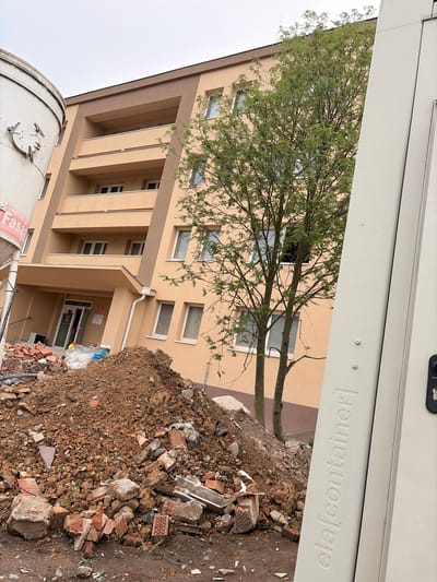 The image depicts a construction site in Příbram, Czechia. In the foreground, a large pile of dirt and debris, including bricks and rubble, is visible. To the left, a portion of a large, cylindrical white container with visible markings and latches is present. To the right, a tall, light-colored shipping container with embossed text reading "ela[container]" is partially visible. Behind the debris pile and the containers, a multi-story building with a peach-colored facade and brown trim stands. Several balconies with beige railings are visible on the building. A tree with green foliage is positioned in front of the building. The sky is overcast. The scene suggests ongoing construction or demolition work. No people are present in the image.