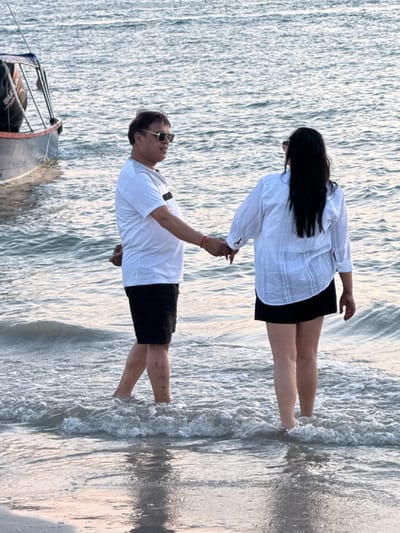 Two individuals, an adult male and an adult female, stand hand-in-hand in the shallow waters of Cenang Beach, Malaysia. The man, positioned on the left and facing partially right, wears sunglasses, a white t-shirt with a gray collar and a black horizontal stripe, and black shorts; his lower legs display multiple dark marks. The woman, positioned on the right with her back predominantly to the viewer, has long dark hair and wears a white striped long-sleeved shirt and a black mini-skirt; she also wears sunglasses, visible above her hair. Both individuals are barefoot, with water reaching approximately mid-calf. To the far left, a small, open boat with a blue hull and a visible "SUZUKI" outboard motor is partially visible, floating in the water further offshore. The water is calm with gentle waves breaking around their feet.