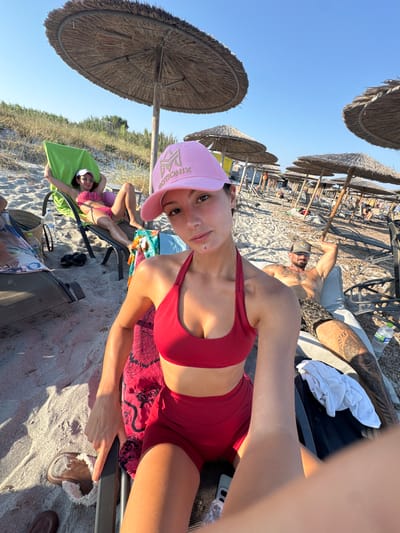 A group of people are relaxing on a sandy beach in Moudros Municipal Unit, Greece, under thatched umbrellas. In the foreground, a woman wearing a red bikini and a pink cap takes a selfie. Behind her, another woman in a pink bikini reclines on a lounge chair. To her right, a tattooed man in patterned shorts is also lounging. Several beach chairs and umbrellas are visible in the background, along with distant figures and sparse vegetation. The weather appears sunny and clear.