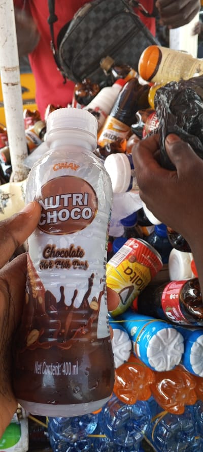 In Awjaw, Nigeria, a close-up photograph captures a retail scene involving beverages. In the foreground, a dark-skinned hand holds a clear plastic bottle of "CWAV NUTRI CHOCO," a 400 ml "Chocolate Malt Milk Drink" exhibiting condensation. The bottle is vertically oriented with its white screw cap visible. In the background, a dense assortment of various bottled and canned drinks is visible, suggesting a vendor's stall. These include dark brown bottles, a yellow and red "MALTA GUINNESS" can, and numerous clear plastic bottles containing blue and orange liquids, many bundled together with yellow elastic bands. Another pair of dark-skinned hands is partially visible on the right, one hand holding a crumpled black plastic bag amidst the other products. A person wearing a red garment and a black checkered bag is partially visible in the upper left background. A white vertical pole or pipe stands on the left.
