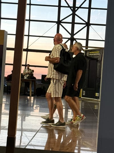 The image depicts two men walking in what appears to be a public indoor space with a large, grid-like window overlooking a sunset. The man in the foreground on the left is bald and wearing a striped shirt and shorts. He has a backpack over his left shoulder. The man on the right is wearing glasses, a black shirt, and black shorts. They are walking side-by-side on a polished floor that reflects the surrounding environment. In the background, a third person is visible behind a table, looking down at something. The overall scene suggests a relaxed atmosphere, possibly an airport lounge or a public concourse, given the large windows and the presence of luggage in the background. The specific location in Zaventem, Belgium, is not explicitly identifiable from the visual information presented.