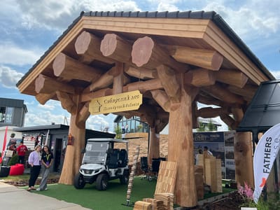 In Yakunino, Russia, this image captures an outdoor exhibition area with a large, rustic wooden structure serving as an entrance or pavilion. The structure is built from massive, rounded logs, with the roof supported by thick log pillars. A sign hanging from the structure reads "Сибирский лес" (Siberian Forest) and "Дома Ручной Работы" (Handmade Houses).
In the foreground, a white golf cart with black seats is parked on artificial green turf. To the left of the cart, two young women are standing and conversing; one wears a light purple top and black pants, while the other is in a black t-shirt and grey pants. Behind them, other exhibition booths and structures are visible, including what appears to be a small building with a dark roof and a large red beanbag chair.
Further in the background, more exhibition structures and attendees can be seen. A large white banner with the text "FATHERS HOUSE" is positioned on the right side of the frame. The environment suggests a sunny day with a partly cloudy sky. The ground is covered with artificial turf in the immediate vicinity of the pavilion and beyond it, with some natural dirt and landscaping visible further back. The overall scene depicts an event showcasing wooden house construction and related products.