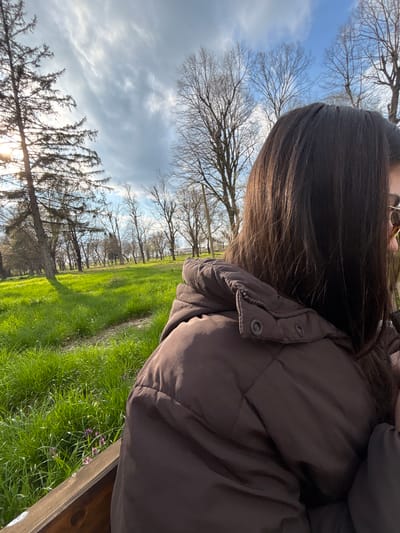 A person is sitting on a wooden bench in a park or grassy area. The person is wearing a brown puffer jacket and has dark brown hair. The view is from behind the person, angled towards the sky and trees. The grass is green, and there are some purple flowers near the bench. Bare trees are visible in the background against a partly cloudy sky. The location is not identifiable in the city of Gromshin, Bulgaria without further information.