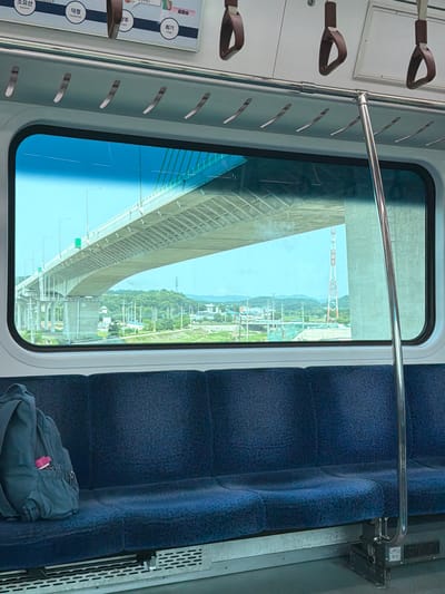 The image is taken from inside a train carriage, looking out of a large window. The seats in the foreground are upholstered in dark blue fabric and appear empty except for a dark blue backpack on the left side of the frame. Above the window, the train ceiling is visible with several brown hand straps hanging down.
Through the window, a large overpass or bridge dominates the view, stretching across the frame. The bridge is supported by concrete pillars. Beyond the bridge, a green, hilly landscape is visible, dotted with trees and some buildings. A tall, lattice-like structure, possibly a transmission tower, stands in the middle ground. The sky is clear and bright blue.
The context provided states the location is Yangju, South Korea. This suggests the bridge and surrounding landscape are part of the scenery visible from a train traveling through or near Yangju. The sign above the window, partially visible in the cropped images, displays Korean text which likely indicates train stations or route information relevant to Yangju.
