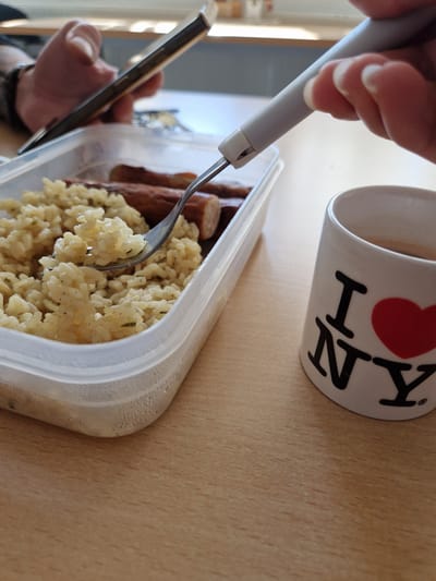 A high-angle shot depicts a meal being eaten on a wood-grain surface. The food, consisting of sausage and rice, is served in a clear plastic container. A silver fork with a white plastic handle is lifting a portion of rice. A hand holding a cell phone is also visible. A white mug, with "I (heart) NY" printed in black and red, sits adjacent to the meal. This scene is likely taking place in Obernburg am Main, Germany.