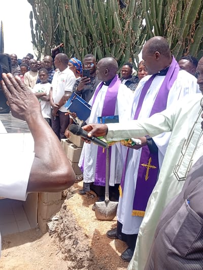 This image depicts an outdoor religious ceremony, likely a burial or a groundbreaking, taking place in Laduran Foron, Nigeria. The scene is set under bright daylight, with a large gathering of people, predominantly men, observing the proceedings.
In the foreground, a person's arm is raised with an open palm, seemingly in a gesture of prayer or blessing. Several Christian clergy members are central to the event, dressed in white robes and purple stoles, some embroidered with gold crosses. One clergyman holds a microphone, while others hold books or binders, including a blue one with partially visible text "God..." and a black one with text that appears to be "Globus" or similar. A shovel rests on the ground beside one of the clergy, implying digging or a ceremonial act.
The ceremony is taking place next to a dug-out area in the reddish-brown earth, which contains stacked concrete blocks and some white tiled surfaces, suggesting either a grave or a foundation for a structure. The numerous onlookers, including men, women, and children, are attentive; some are holding mobile phones, presumably recording the event.
The background is characterized by a dense cluster of tall, columnar cacti, indicating a dry or semi-arid environment. The overall atmosphere appears solemn and respectful, reflecting a significant community or religious ritual.