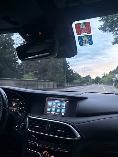 The image shows the interior of a car parked on a street, likely in Brugg, Switzerland, given the presence of Swiss vignette stickers on the windshield. The view is from the driver's perspective, looking forward through the windshield and at the dashboard. The dashboard features a digital display showing the time as 21:19 and a temperature of 18.9°C. The speedometer shows a reading of 48. The car's central infotainment screen displays a phone interface with options like "Favorites," "Call History," and "Phone Book."
Outside the car, the street is visible, lined with trees and buildings. The sky is overcast, suggesting it is late evening or dusk. The overall scene depicts a common urban environment, possibly during a stop or while waiting. There are no people visible inside or outside the car.