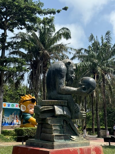 In Nanwan Village, China, a statue of a chimpanzee is depicted contemplating a sphere, resting on a stack of books inscribed with the name "DARWIN". To the left, a yellow cartoonish monkey figure wearing sunglasses and a green shirt sits in front of a colorful mural depicting ocean waves and cartoon characters. The scene is set outdoors with lush palm trees and other foliage under a bright, partly cloudy sky. A few people are visible in the background, walking on a paved path, and a red-brick pedestal supports the main statue.