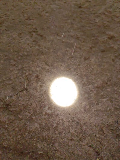 A close-up, slightly out-of-focus shot shows a textured surface, possibly earth or ground cover, with a circular, bright light source in the center. There are what appear to be small pieces of debris or plant matter scattered across the surface, and possible raindrops in the upper portion of the image. The exact location within Slatina, Bulgaria, cannot be determined from the provided image. No people or other subjects are visible.