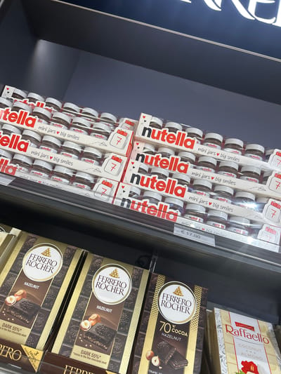 The image shows a display of various sweets. On the left, stacked packages of Nutella "mini jars, big smiles" packs are visible. To the right, there are boxes of Ferrero Rocher chocolates and a box of Raffaello coconut and almond confectionery. The picture likely captures a scene from a retail store within Nouaceur, Morocco.
