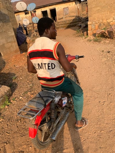 A man lies on his side on a red motorcycle that is on its side in the dirt. He wears green pants, sandals, and a white shirt with the word "LIMITED" printed on it. Beside the motorcycle are bottles and containers secured with a net. In the background, there is a brick building and a dirt path, along with satellite dishes mounted on the wall.