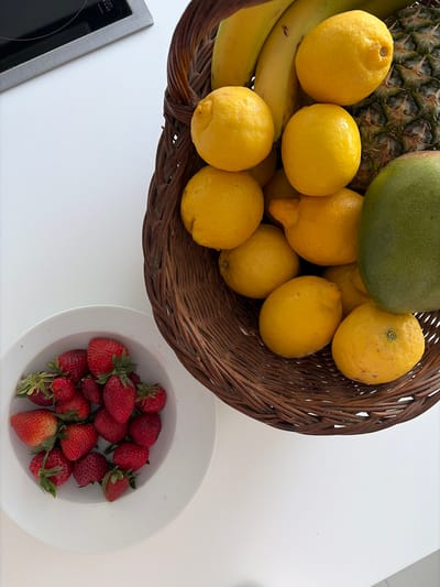 A high-angle shot depicts a white tabletop with a wicker basket filled with fruit and a white bowl of strawberries. The basket contains bananas, lemons, a pineapple, and a green mango. The bowl of strawberries is positioned to the right of the basket. A portion of a built-in stovetop is visible in the lower left corner. The image does not provide enough information to determine a specific location within the Municipal Unit of Elliniko, Greece.