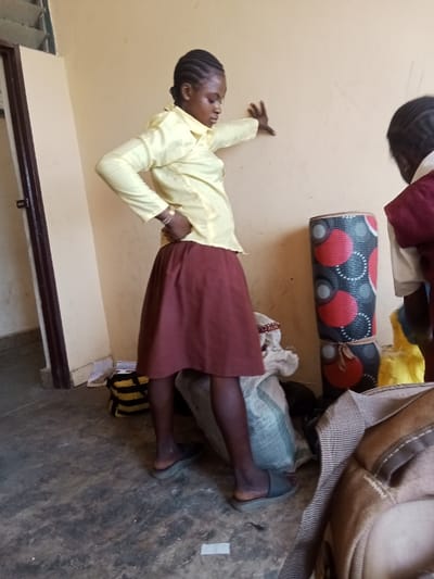 An indoor scene in Gidan Kado, Nigeria, depicts a female individual standing with her left hand against a light-colored wall and her right hand on her hip, looking downward. She wears a light yellow long-sleeved shirt, a dark red knee-length skirt, dark hosiery, and grey slide sandals. A gold-colored bracelet is visible on her left wrist. Her hair is styled in braids. To her right, the upper back and shoulder of a second individual, wearing a white shirt and a dark red garment, are partially visible. A rolled-up mat with black, red, and white circular patterns stands vertically against the wall. Various items are on the floor, including a light-colored sack, a black and yellow striped bag, scattered papers, and a partially visible light brown shoulder bag in the foreground. A dark door frame is present on the left side of the image, revealing an opening to another area, with a horizontal-slatted window or vent at the top left. The floor is dark grey concrete with light patches and debris. The walls are solid and light-colored.