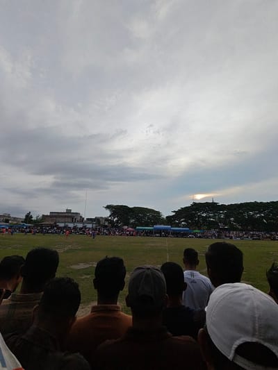 In the city of Desa Meunasah Blang, Indonesia, a soccer match is taking place on a grassy field. A large crowd of spectators is gathered around the perimeter of the field, some under tents. The sky is overcast, but the sun is visible through a break in the clouds. In the foreground, the backs of several spectators are visible, facing the field. The spectators are dressed in casual clothing. The scene depicts a typical community sporting event.