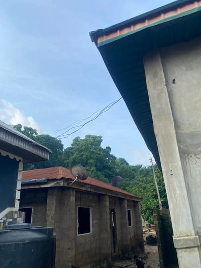 The image displays a daytime scene in Igarra, Nigeria, characterized by residential structures and dense vegetation under a light blue sky with scattered clouds. In the foreground on the right, a large, unpainted concrete building features a dark green-eaved roof extending over a rough concrete wall and a prominent vertical concrete column. Several thin, dark wires, likely power lines, traverse the sky horizontally across the middle of the frame. To the midground left, a building with a light gray corrugated roof and decorative fascia board is partially visible, with a black plastic water tank occupying the lower-left corner. Central to the midground is a smaller, single-story structure constructed from concrete blocks, exhibiting a rusty-red corrugated metal roof. This building has multiple window openings, some framed in white, and a small, weathered satellite dish is affixed to its roof. The background is dominated by a dense canopy of green trees and foliage, interspersed with large rock formations visible through the greenery. A dirt path separates the midground buildings, and a dark animal, possibly a goat, is partially visible on this path in the lower right. A wooden pole is also present amidst the vegetation in the midground right.