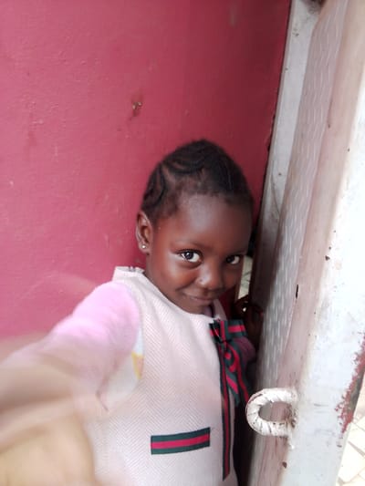 A young Black girl with braided hair and a white vest over a pink shirt, adorned with a ribbon featuring green and red stripes, stands outdoors in front of a pink wall and a white, textured metal structure. The girl is looking towards the camera with a slight smile. The setting appears to be an urban environment, possibly Mbar, Nigeria, given the textured concrete or stucco of the wall and the weathered metal, which could be part of a building facade or a street fixture. The lighting suggests it's daytime, with natural light illuminating the scene. The girl's left hand is blurred in the foreground, indicating movement or the camera's focus.