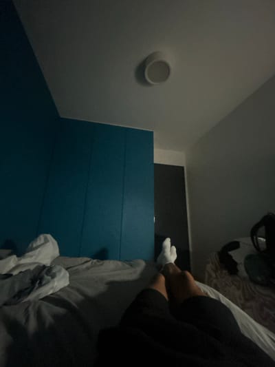 The image depicts an indoor scene, likely a bedroom, with a person lying on a bed. The person's legs and feet, clad in white socks, are visible in the foreground, with dark pants covering the rest of their legs. The bed appears to be made with grey sheets. To the left of the frame, a wall painted in a vibrant teal color dominates the space. Above, a white circular light fixture is recessed into the ceiling. To the right, a portion of a doorway or a closet is visible, along with what appears to be a backpack or bag. The lighting suggests it is either nighttime or the room is dimly lit. The location is stated to be in Carrières-sous-Poissy, France.