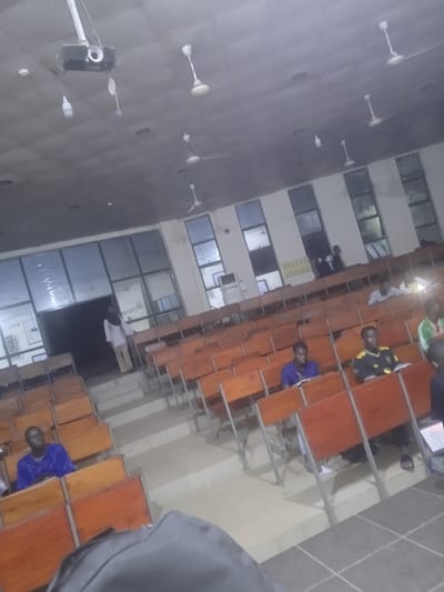 The image shows an interior view of a lecture hall in the city of Ife, Nigeria. Several rows of orange wooden chairs with metal frames are visible, arranged in tiered seating. In the foreground, a person's backpack is partially visible. Several individuals are seated in the chairs, appearing to be students. One person in the middle row is wearing a blue shirt. Another person in the row behind them is wearing a black and yellow jersey. Further back, other individuals are seated, some with books or papers in front of them. At the entrance to the hall, a person wearing a white shirt and headscarf is standing. Above, the ceiling is equipped with multiple ceiling fans and a projector is mounted, along with a light bulb. The lighting in the hall appears to be artificial.