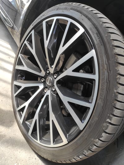 The image shows a close-up of a car wheel. The wheel has a black and silver multi-spoke design. The tire is a Hankook Ventus Evo SUV. The tire has some scuff marks on the tread. The car appears to be parked on a concrete surface, possibly in an outdoor parking lot. The background is blurred, but suggests a slightly overcast day. The context of the image is a photograph taken in Saint-Genis-Laval, France, with the car identified as a Volvo XC40.