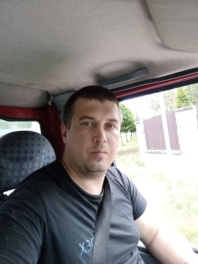 The media file is a close-up selfie taken from inside a vehicle, likely a van or truck, looking forward. The primary subject is a man, appearing to be in his late 20s to 30s, with short brown hair and a light beard. He is wearing a black t-shirt with a light blue graphic that appears to read "EX" or "MEX" on the left side of his chest. The man is wearing a seatbelt, indicating he is either driving or is a passenger in a moving vehicle.
The interior of the vehicle is visible, including the grey headliner, a red trim piece above the window, and the textured fabric of the passenger seat to the left. Outside the window, a fence with vertical wooden planks and stone pillars can be seen, along with trees and overgrown grass. The lighting suggests it is daytime, with bright outdoor light filtering into the vehicle. The overall impression is a casual, candid shot, possibly taken while traveling.
The location context provided is Svémyslice, Czechia. This information, combined with the visual cues of the fence and vegetation, suggests a rural or suburban setting in the Czech Republic. There are no strong indicators of a specific emotion, beyond a neutral or slightly contemplative expression on the man's face.