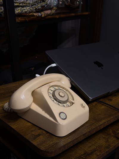 The image shows a beige rotary dial telephone resting on a dark, wood-grained surface. A silver laptop, potentially a MacBook based on the visible Apple logo, is positioned adjacent to the phone. A white USB cable connects the laptop and the phone. The image is taken in a dimly lit interior setting with a partially visible rack with clothes. There are no people visible in the frame. The phone and laptop are the primary objects of focus. There is no indication of the location in the city of Yeysk, Russia.