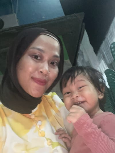 In Wedarijaksa, Indonesia, a medium shot captures a woman and a young child posing for a selfie. The woman, wearing a dark hijab and a yellow and white patterned shirt with buttons, is on the left, looking at the camera with a slight smile. Her makeup includes lipstick. To her right, a child in a pink long-sleeved shirt is smiling widely, with their hand near their mouth. The background is dimly lit and appears to be outdoors or in an open area, with hints of structures and possibly furniture. The overall impression is a casual, candid moment shared between the two individuals.
