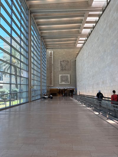 The image depicts the interior of a modern airport terminal in Tzafria, Israel. The scene is characterized by a vast, open space with high ceilings supported by concrete beams. The left side of the terminal features a floor-to-ceiling glass facade, offering a view of palm trees and greenery outside. The right side is dominated by a large, textured stone wall.
In the mid-ground, a moving walkway is visible, with two individuals, one wearing a red top and the other in a black top with a headscarf, standing on it. Further down the walkway, more people are seen, along with a small white utility vehicle. The floor is made of polished stone tiles.
On the stone wall, three large art installations are displayed. The two lower ones are square mosaics, while the uppermost one is a long, vertical rectangular tapestry. Above the entrance to a passage, a sign reads "WELCOME" in English, accompanied by Hebrew text.
The lighting in the terminal is a combination of natural light streaming through the glass windows and artificial light from the ceiling. The overall atmosphere is one of spaciousness and modern design.