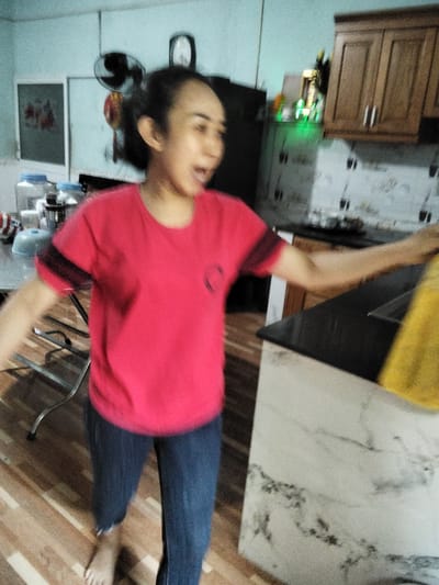 The image depicts a person in what appears to be a kitchen in Phú Sơn, Vietnam. The individual, wearing a red t-shirt and blue jeans, is captured mid-action, with their mouth open as if speaking or reacting. They are standing on a wooden floor, and their right arm is extended, holding a yellow cloth. To the left, a table with various containers, including plastic bottles with blue lids, is visible. In the background, kitchen cabinets with dark wood doors are mounted on the wall. A fan is also mounted on the wall above and behind the person, with a partially obscured clock above it. The environment suggests a residential kitchen setting, likely in Phú Sơn.