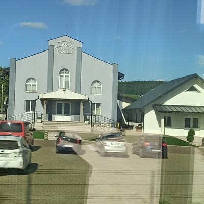 The image shows a modern, light-gray church building with arched windows and a prominent sign reading "BISERICA PENTICOSTALA DANILA". The church has a large entrance with double doors and a small porch area. To the right of the main church building is a smaller, white building with a dark roof and a triangular window. Several cars are parked in the paved lot in front of the buildings. A red van is parked to the left, and several sedans are parked in the center and right side of the image. The buildings are set against a backdrop of green trees and hills under a clear blue sky with a few wispy clouds. The location appears to be in Dănila, Romania, as indicated by the church's signage.