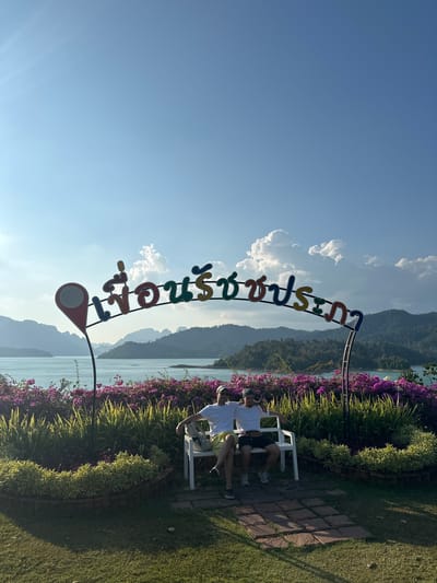 Two people are sitting on a white bench in Ban Chiao Lan, Thailand, near a body of water and mountains. A colorful sign in Thai script stands in front of them. The area is landscaped with flowers and grass.
