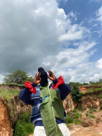 The image captures a person standing in a rugged, outdoor landscape with a dramatic sky overhead. The person is wearing a blue and white striped sweater, a black beanie, and white pants. They are holding a large green leaf in front of them and have their hands raised, fingers forming a gesture. The background features a steep, eroded embankment with exposed reddish-brown earth and green vegetation. The sky is partly cloudy, with areas of bright blue interspersed with large, dark gray clouds, suggesting changing weather. The overall scene is set in a natural, possibly remote location within the city of Ufu Esioni, Nigeria.