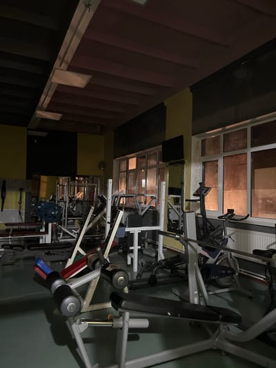 The image depicts the interior of a gym in Kadamjay, Kyrgyzstan. The room is filled with various weightlifting and cardio equipment, including benches, weight racks, dumbbells, and exercise bikes. The lighting is dim, with fluorescent lights visible on the ceiling and light filtering in from the windows. The walls are painted in a combination of yellow and dark colors, and there are radiators beneath the windows. The overall scene suggests a functional but somewhat dated fitness facility.